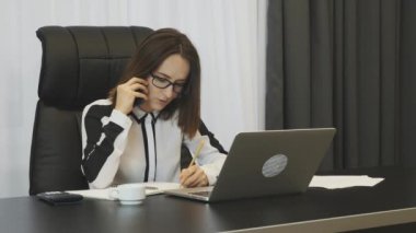 Businesswoman working in modern office. Confident female writing notes while talking on phone. Thoughtful concerned woman negotiates by phone at her office. Employee works on laptop. Business concept
