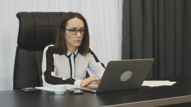 Female works on laptop in office. Young concentrated woman in business clothes working on laptop computer and taking notes in notebook. Office employee doing job. Brunette businesswoman keeps account
