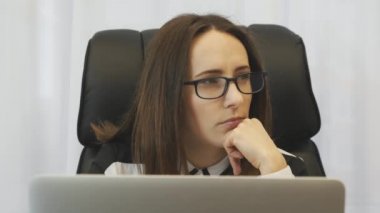 Businesswoman thinking about her work in office. Young concentrated woman in white blouse working on laptop in modern office. Portrait of confident female focused on her job