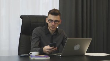 Modern ofiste çalışan bir iş adamı. Odaklanmış genç adam iş yerinde çalışıyor. Ofis masasında oturan bir erkek telefon ekranına bakıyor. Kendine güvenen bir çalışan işini yapıyor. Akıllı telefonda yazan adam.