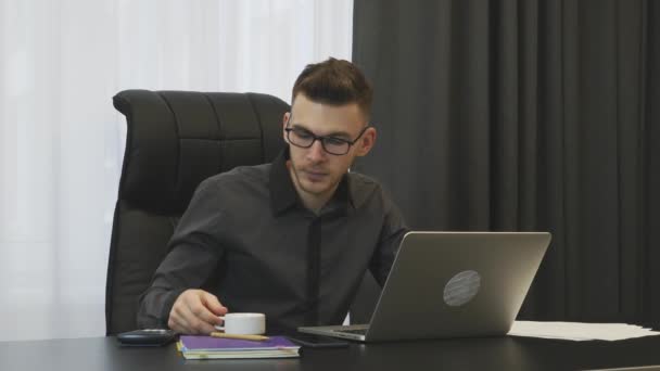 商人在工作场所喝咖啡浓缩咖啡.年轻自信的戴眼镜的男人坐在他现代的办公室里喝咖啡。男性办公室职员在笔记本电脑上工作，喝咖啡 — 图库视频影像