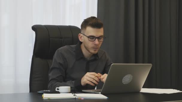 Takım elbiseli ve gözlüklü bir iş adamı modern ofisinde çalışıyor. Başarılı bir adam iş yerinde çalışır. Erkek danışman dizüstü bilgisayara bakıyor ve not alıyor. Finansör şirketin hesaplarını tutar — Stok video