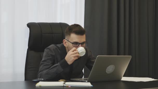 Egy férfi kávét iszik a modern irodájában. Fiatal üzletember öltönyben és szemüvegben kávét iszik a munkahelyén. Férfi pénztáros, aki az irodájában ül és eszpresszót kortyolgat. Üzleti sikerkoncepció — Stock videók