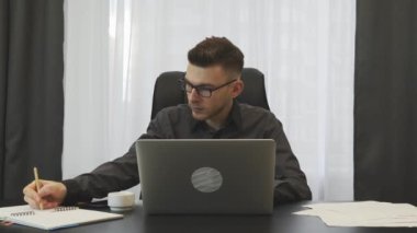 Male financier working on laptop in office. Young successful businessman works at workplace and takes notes in notebook. Confident focused man keeps financial report. Business victory concept
