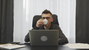 Successful businessman drinking coffee in his modern office. Young man in suit drinks coffee at workplace. Male broker sitting at his office desk and sipping espresso. Business success concept