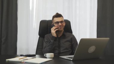 Successful young businessman talking on phone in his modern office. Man talks with partners on smartphone and makes deal. Male financier speaking on phone sitting at his office desk. Business concept