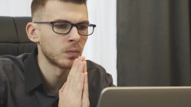 Laptop ekranına umutla bakan bir adamın portresi. İşadamı bilgisayar ekranına bakarak dua ediyor. Ofis masasında oturan genç bir erkeğin yakın görüntüsü. İş başarısı konsepti
