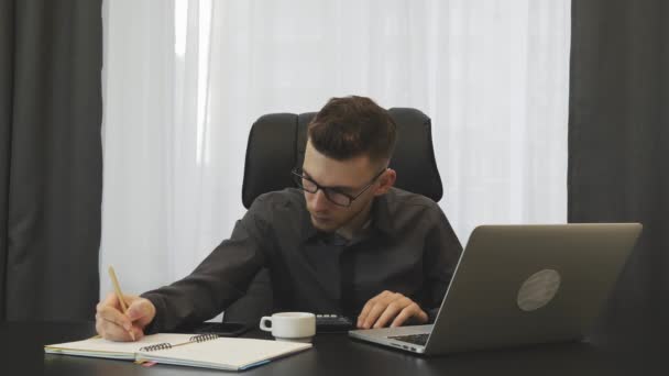 Businessman keeps accounts in modern office with laptop. Young male accountant checking financial report. Young man makes report using calculator and notebook. Broker working at workplace in office — Stok video