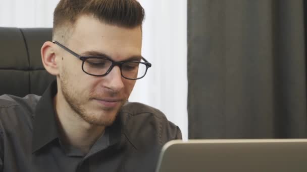 Primer plano del hombre que trabaja en el ordenador portátil en la oficina. Retrato de hombre de negocios en gafas mirando la pantalla de la computadora. Joven hombre confiado trabaja en el ordenador portátil en la oficina. Hombre leyendo buenas noticias en internet — Vídeos de Stock