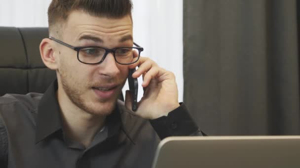 Muž mluví po telefonu v kanceláři, zblízka. Úspěšný obchodník s brýlemi mluví po telefonu a dívá se na monitor notebooku. Portrét usmívajícího se muže mluvícího na smartphonu s klienty v kanceláři — Stock video