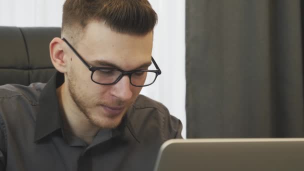 En mann som feirer gode nyheter på nettet. Portrett av forretningsmann begeistret med vellykket jobbforfremmelse sittende på arbeidsplassen. Ung smilende mann som leser positive nyheter på PC-skjermen – stockvideo