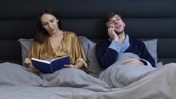 Mujer y hombre acostados en la cama antes de dormir. Familia joven pareja se encuentra en el dormitorio. Mujer leyendo libro y hombre hablando por teléfono en la cama. Una joven pareja casada descansando en la cama. Relación romántica — Vídeo de stock