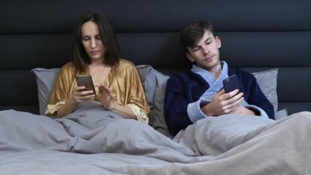 Manželský pár v posteli hledající smartphony. Rodinný pár používající pomůcky leží na posteli, pár závislý na technologii. Žena a muž používající gadgets ležící na šedé posteli. Moderní rodina o víkendu doma — Stock video