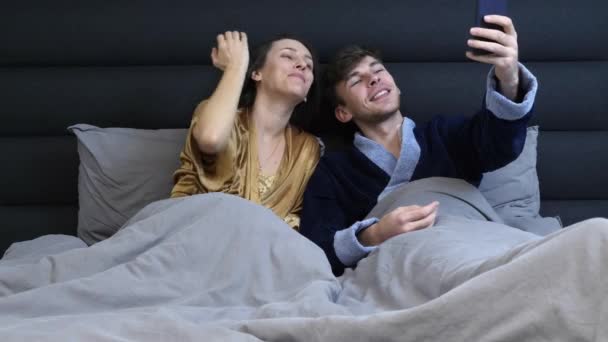 Familia moderna tomando selfie en el teléfono inteligente acostado en la cama gris. Una joven pareja casada tomando selfie en la cama del hotel. Divertido hombre y mujer en la cama cómoda en la habitación de hotel tomando una selfie por la mañana — Vídeos de Stock