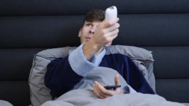 Genç erkek klimayı uzaktan kumandayla açıyor, portre kullanıyor. Yatakta bornozlu bir adam uzaktan kumandalı düğmelere basıyor. Otel odasında yatakta yatan adama yakın dur.