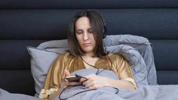 Mujer joven dormida en ropa interior nocturna acostada en la cama y escuchando música en los auriculares. Mujer molesta en los auriculares grandes escucha la música se encuentra en el dormitorio del hotel. Concepto de confort doméstico — Vídeos de Stock