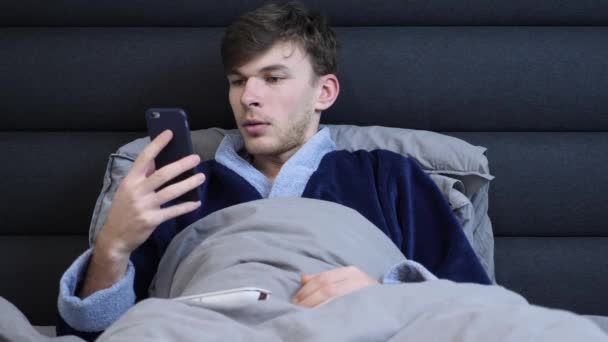 Молодой человек в халате лежит в постели и читает новости на смартфоне перед сном. Мужчина лежит в постели и смотрит на экран телефона утром. Белый парень в постели в гостиничном номере — стоковое видео