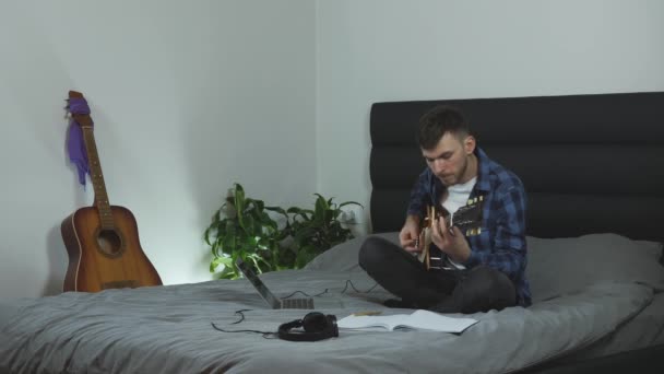 Гітарист грає на гітарі вдома на ліжку. Музикант у навушниках грає на електрогітарі. Молодий чоловічий тисячоліття практикує музичний інструмент. Концепція музики — стокове відео