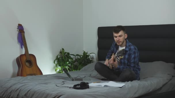 ギターで演奏する若い千年。モダンなリビングルームのベッドに座っているエレキギターでパントンブルーのTシャツの魅力的な男が演奏します。男性音楽家、ギター練習。男演劇ギター-ホーム — ストック動画