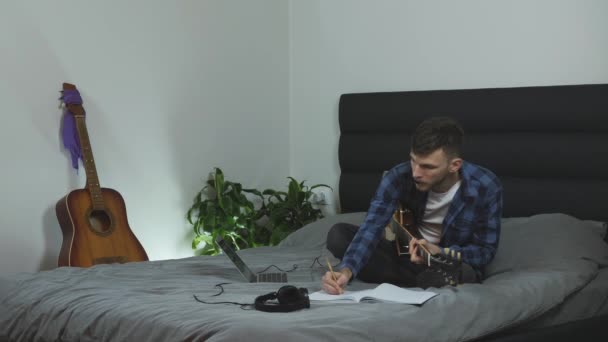 Man övar gitarr. Ung kaukasier man i pantone blå t-shirt är att lära sig på elgitarr på sängen i moderna hem. Musiker skapar ny sång hemma. Attraktiv kille som spelar på gitarr — Stockvideo