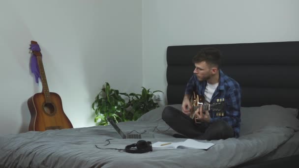 O homem toca rock na guitarra. Jovem músico tocando guitarra elétrica sentado na cama na moderna sala de estar. Homem a praticar guitarra. Emocional millennial executando sua nova música. Conceito musical — Vídeo de Stock