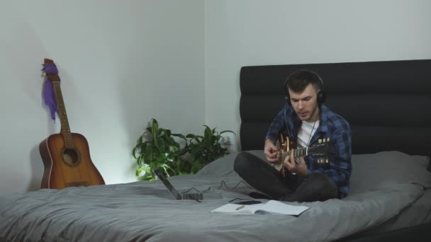 Homem em fones de ouvido cantando e tocando guitarra elétrica. O músico masculino canta sua nova canção na cama na moderna sala de estar. Guitarrista a praticar aulas de guitarra. Millennial está cantando música romântica — Vídeo de Stock