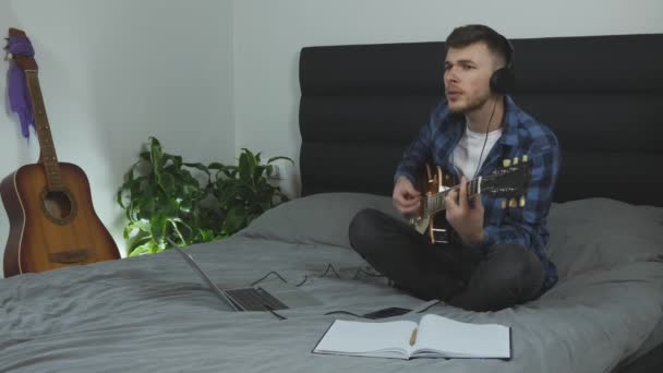 Homem emocionalmente cantando e tocando guitarra elétrica. Jovem bonitão toca guitarra em casa. Músico aprendendo música nova. Guy em fones de ouvido executa música lírica na cama na sala de estar moderna — Vídeo de Stock