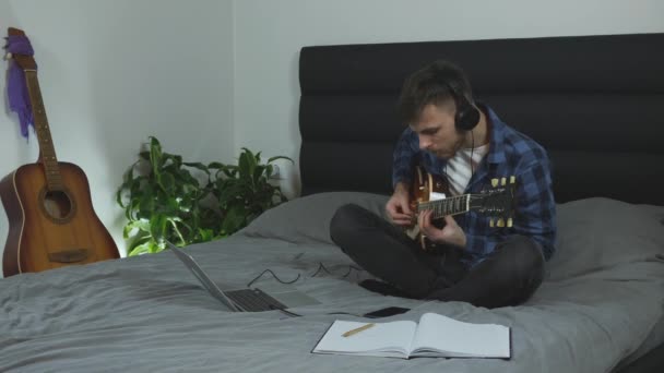 Een man die gitaar oefent. Gitarist leren nieuwe akkoorden op elektrische gitaar op bed in het moderne huis. Muzikant in hoofdtelefoon die romantische lyrische muziek maakt. Gitaarlessen. Muziekconcept — Stockvideo