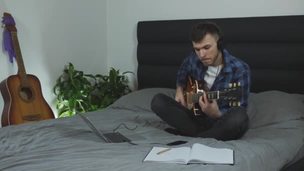 Gitarrist i hörlurar spelar på elgitarr. Musiker som spelar gitarr på sängen i vardagsrummet. En ung kaukasisk man i pantonblå t-shirt som övar på sin nya sång. Gitarrläraren sjunger sång — Stockvideo
