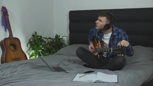 Profesorul de chitară cântă corzi de chitară. Tânăr în căști tuning chitară electrică pe pat la domiciliu. Muzician de sex masculin cu chitară acasă. Conceptul muzical — Videoclip de stoc