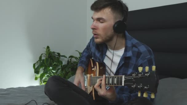 Retrato de un joven milenario tocando la guitarra eléctrica. Guitarrista interpreta música lírica. El hombre de los auriculares toca la guitarra. Guapo caucásico chico practicando nuevos acordes en la cama en casa — Vídeos de Stock