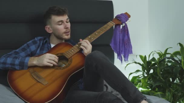 Joven guapo barbudo músico tocando rock pop emocional en la guitarra acústica y cantando sentado en la cama . — Vídeo de stock