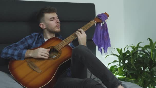 Joven músico barbudo tocando emocionalmente la canción de balada rock en la guitarra acústica en la cama en casa . — Vídeo de stock