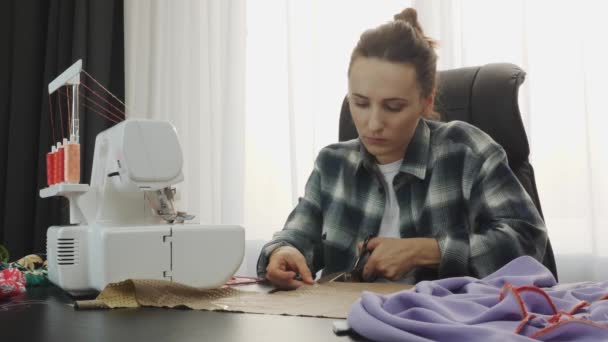 Terzinin stüdyosundaki masada makas ve kumaşla çalışan bir kadın. Kadın el yapımı elbiseyle çalışıyor. Kumaş dikme atölyesi. Kadın giyim tasarımcısı atölyede kumaştan elbiseler yapıyor. — Stok video