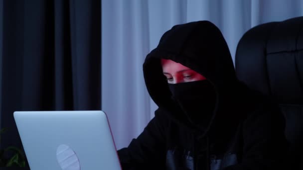 Cara femenina en máscara negra y sudadera con capucha mirando la pantalla del portátil. Cara de hacker en capucha negra mirando el ordenador y tecleando el código. Mujer sentada en el portátil y programación en el teclado. Concepto web de seguridad — Vídeo de stock