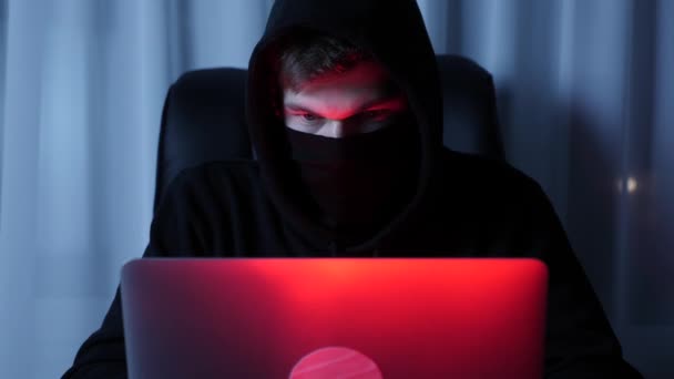 Primo piano di hacker maschile guardando lo schermo del computer portatile seduto nella stanza buia. Uomo in maschera nera e cappuccio digitando sulla tastiera del computer portatile. Faccia da hacker professionista guardando laptop e programmando script di sicurezza — Video Stock