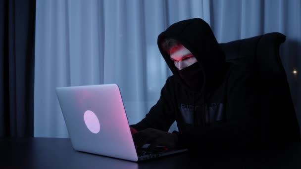 Zelfverzekerde mannelijke hacker op zoek naar laptop scherm. Man met zwart masker en kap zittend in de donkere kamer en het hacken van online sociale bescherming. Jonge hacker breekt computerwachtwoord. Langzame beweging — Stockvideo