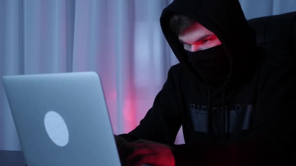 Хакер в черной маске и капюшоне смотрит на экран ноутбука, сидящего в темной комнате. Мужчина в черной толстовке кодирует и программирует на экране компьютера. Видеозапись с поглаживающими клавишами хакера. Медленное движение — стоковое видео