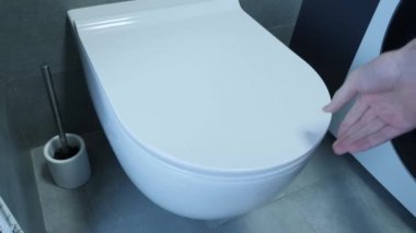 Erkek eli klozet kapağını kaldırır, kapat. Erkekler tuvaletin kapağını duvarlarında ve zemininde gri fayanslarla kaldırıyor. Beyaz tuvaleti, tuvalet fırçası ve çamaşır makinesi olan banyonun yan görüntüsü