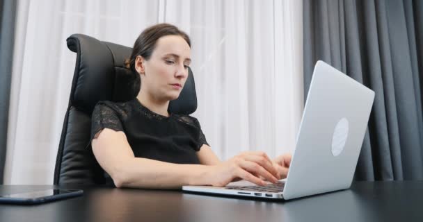 Женщина, работающая с ноутбуком в офисе. Женщина печатает на компьютерной клавиатуре. Леди-босс в черном платье сидит за рабочим столом — стоковое видео