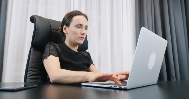 Портрет молодой женщины, работающей на ноутбуке и печатающей на клавиатуре. Уверенная деловая женщина в черном платье сидит в своем офисе. женщина смотрит на экран компьютера — стоковое видео