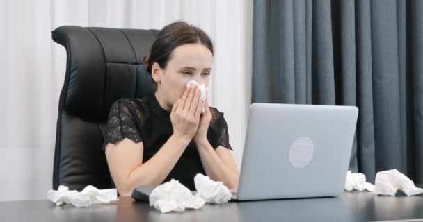 Больная женщина с насморком работает в офисе с грязными бумажными салфетками. Женщина сморкается. Женщина с болезнью и красными глазами сидит за рабочим столом и смотрит на ноутбук — стоковое видео