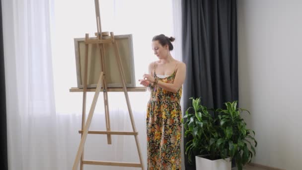 Художня жінка стоїть біля мольберта і малює на полотні, використовуючи акрилові фарби в художній студії — стокове відео