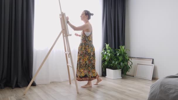 Талантливая молодая девушка с кистью в руке, рисующая картину на холсте. Женщина-художник в домашней студии стоит возле мольберта и рисует на холсте. Процесс рисования. Вдохновение — стоковое видео
