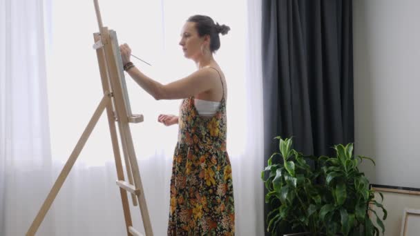 Utalentowana młoda artystka stojąca blisko sztalugi ze szczotką w dłoni i malująca na płótnie. Piękna kobieta w pracowni artystycznej tworzy dzieło sztuki. Inspiracja — Wideo stockowe