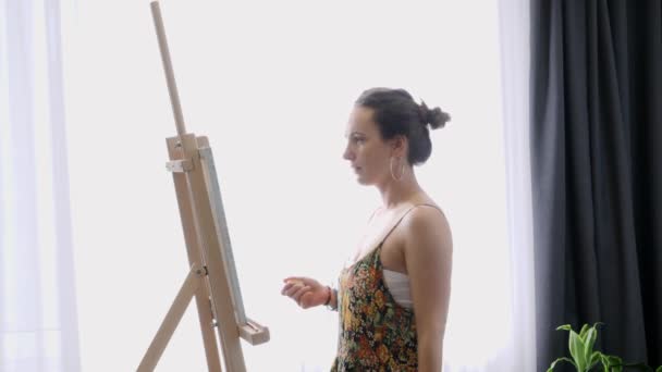 Chica joven inspirada con pincel delgado en pinturas a mano sobre lienzo en taller de arte. La artista femenina está creando pintura abstracta sobre caballete. Academia de arte o escuela de dibujo — Vídeo de stock