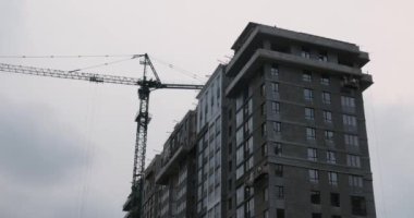 Yüksek vinçli inşaat alanı. İnşaatçılar modern konut kompleksi inşa ediyorlar. Crane, gri bulutlu gökyüzünün altında inşaat alanında çalışıyor. Gökdelen büyük bir vinçle yapım aşamasında.