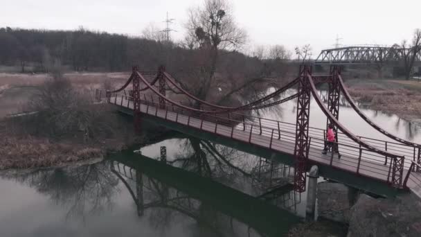 Feminino corre através da ponte pedonal perto do passeio com parque. Mulher correndo ao ar livre, vista aérea. Atleta fazendo exercícios no parque com rio e cais — Vídeo de Stock