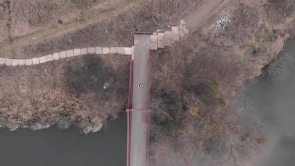 Vista aérea de la mujer corriendo por el puente. Deportiva atleta con chaqueta brillante corre en el puente a través del río en el parque de la ciudad. Motivado joven corriendo al aire libre — Vídeo de stock