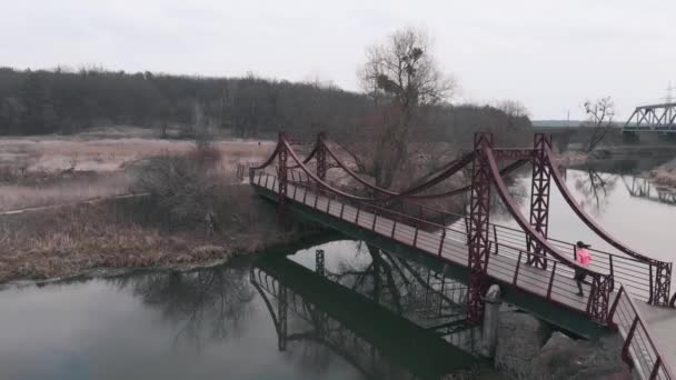 Жінка біжить по мосту через річку, вид з повітряного безпілотника. Брюнетка спортсменка біжить в парку з річкою і набережною. Дівчина практикує бігові вправи в зоні відпочинку — стокове відео